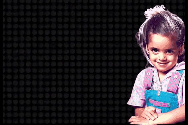 a little girl wearing overalls and a pink shirt with polka dots smiles for the camera