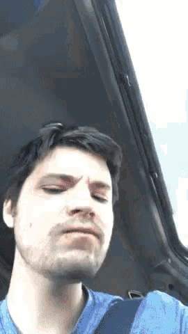 a man with a beard is wearing a blue shirt and a seat belt while sitting in a car .