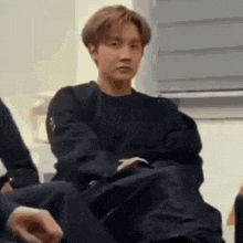 a man in a black sweater is sitting on a couch with his legs crossed .