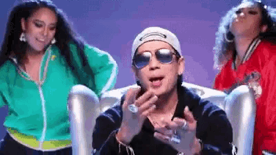 a man wearing sunglasses and a hat is sitting in a chair while two women dance behind him .