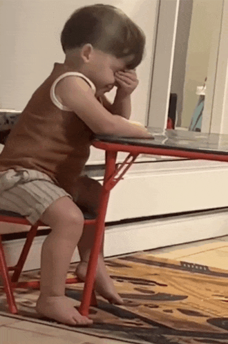 a little boy sits at a table covering his face with his hands