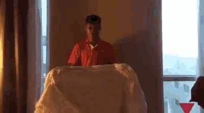 a man in a red shirt is standing in front of a window holding a white blanket .