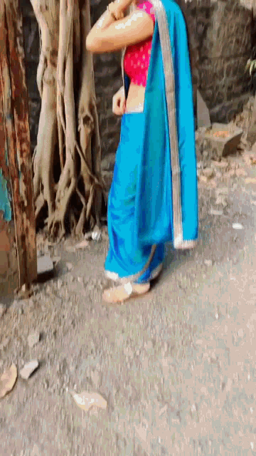 a woman in a blue saree and a red blouse is standing on a dirt road .