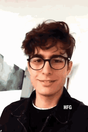 a young man wearing glasses and a black jacket is smiling and looking at the camera .