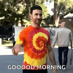 a man wearing a tie dye shirt is walking down a street and says good morning .