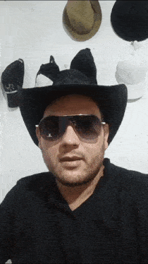 a man wearing a cowboy hat and sunglasses stands in front of a wall full of hats