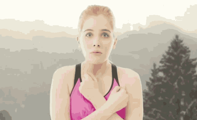 a woman in a pink tank top is standing in front of a mountain and covering her chest with her hands .
