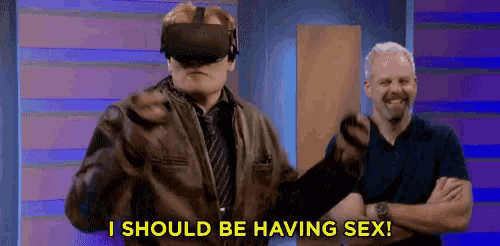 a man wearing a virtual reality headset is pointing at the camera while another man stands behind him .