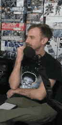 a man in a black shirt is talking into a microphone while sitting in front of a wall of toy boxes