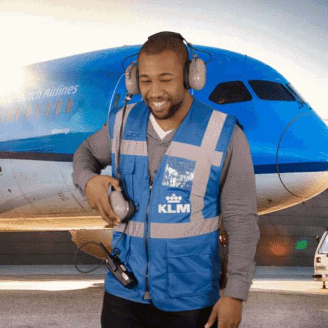 a man wearing headphones and a klm vest stands in front of an airplane