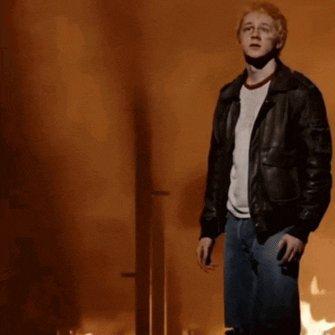 a man in a leather jacket and jeans is standing in front of a wall that is on fire .