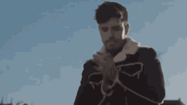 a man with a beard is smoking a cigarette while standing in front of a blue sky .