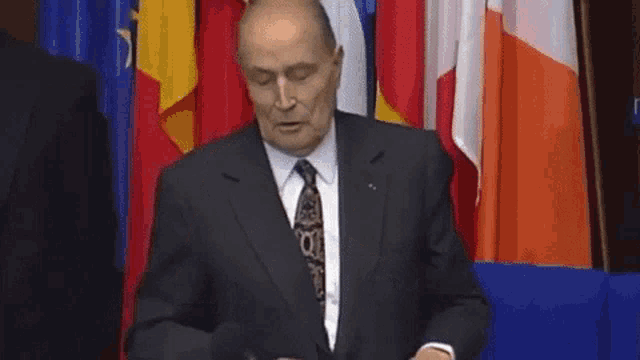 a man in a suit and tie is standing in front of flags and says le nationalisme c'est la guerre .