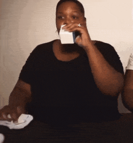 a woman in a black shirt is sitting at a table drinking from a white cup