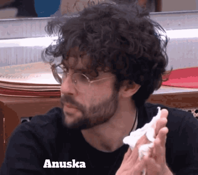 a man with glasses and a beard is holding a napkin and the name anuska is on the bottom right