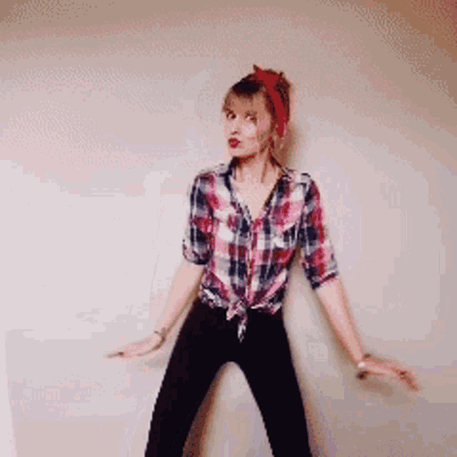 a woman wearing a plaid shirt and black pants is dancing in front of a white wall