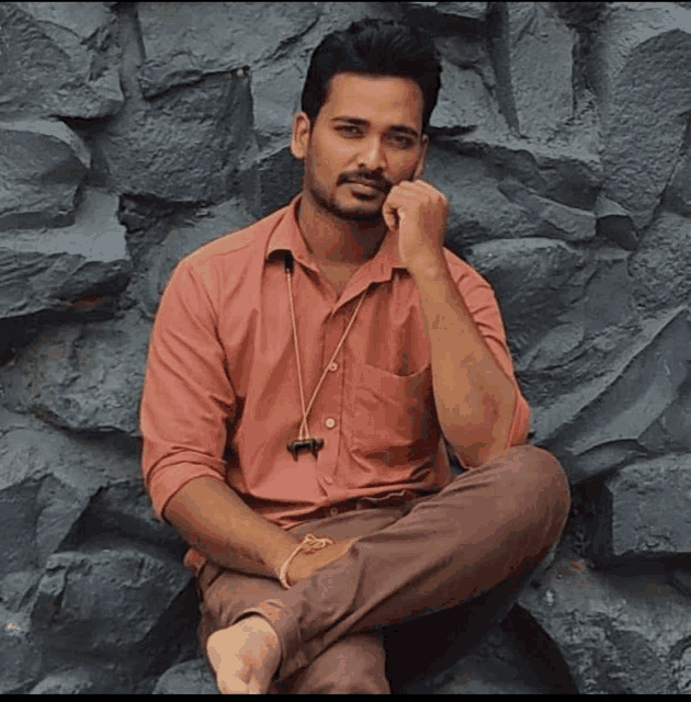a man in a red shirt sits with his legs crossed in front of a stone wall