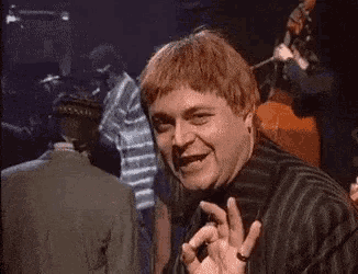 a man with red hair is smiling and making an ok sign in a crowded room .