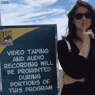 a woman holds her finger to her lips in front of a sign that says video taping and audio recording will be prohibited