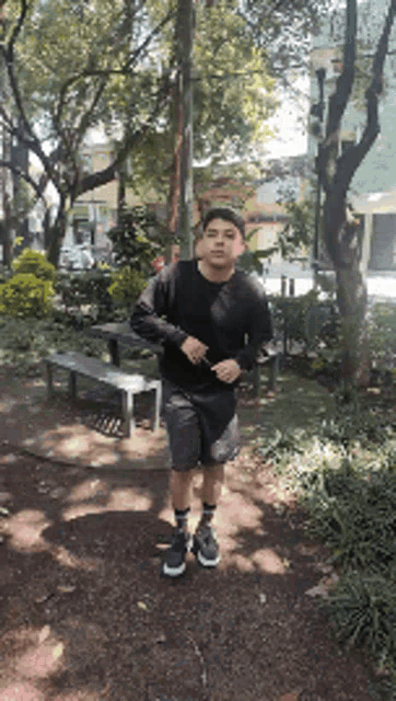 a young man in a black shirt and shorts is standing on a sidewalk in a park