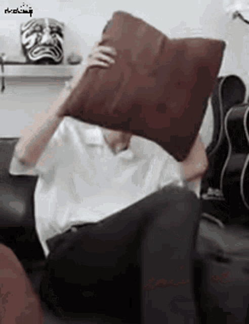 a man is sitting on a couch holding a brown pillow over his head .