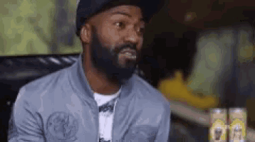 a man with a beard wearing a hat and a jacket is sitting on a couch .