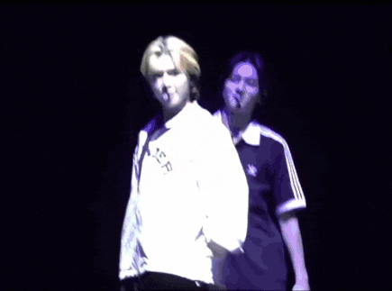 a group of people are dancing in a dark room . one of the people is wearing a purple adidas shirt .