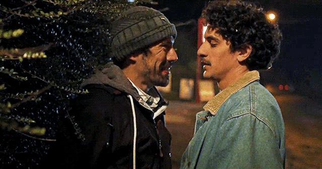 a man wearing a beanie talks to another man in a denim jacket