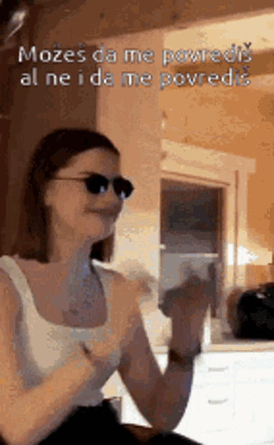 a woman wearing sunglasses and a white tank top is clapping her hands in a room .
