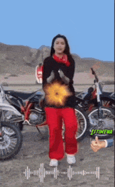 a woman in red pants and a black shirt is standing in front of a motorcycle .