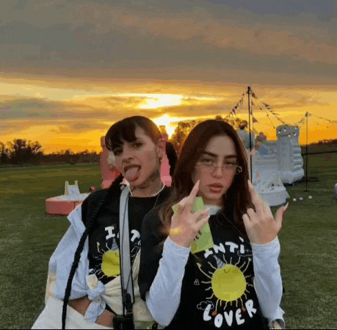 two girls are standing next to each other in a field at sunset and one of them is sticking her tongue out .