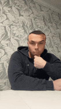 a man sitting at a table with his hand on his chin in front of a floral wallpaper