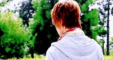 a man with red hair is standing in a field with trees in the background looking at something .