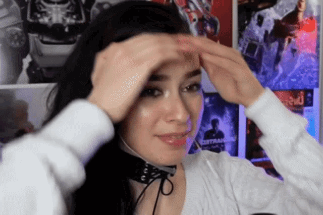 a woman wearing headphones and a choker adjusts her hair in front of a wall with posters on it