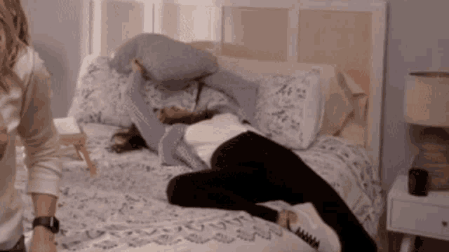 a woman is laying on a bed with a pillow on her head and another woman is standing next to her .