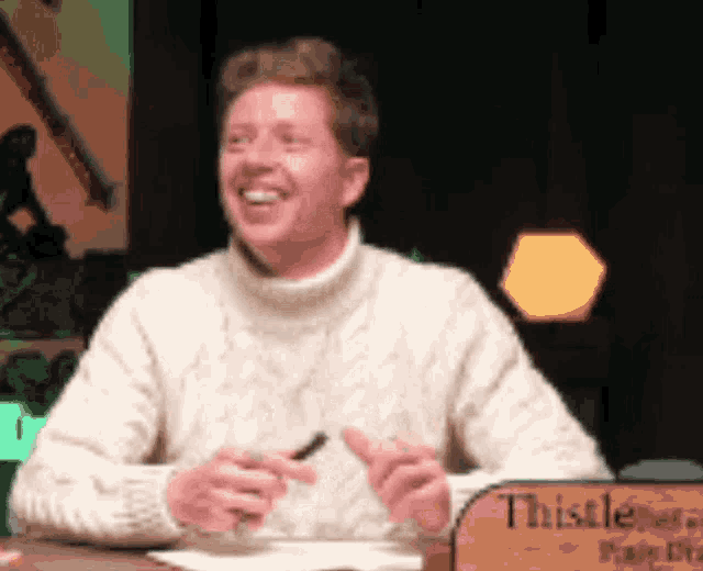 a man in a white sweater is sitting at a table with a sign that says thistle on it