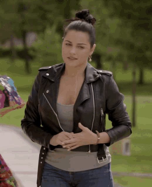 a woman wearing a black leather jacket and jeans stands in a park