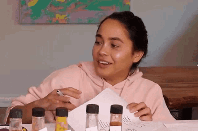 a woman in a pink hoodie is sitting at a table with bottles of spices and a piece of paper .