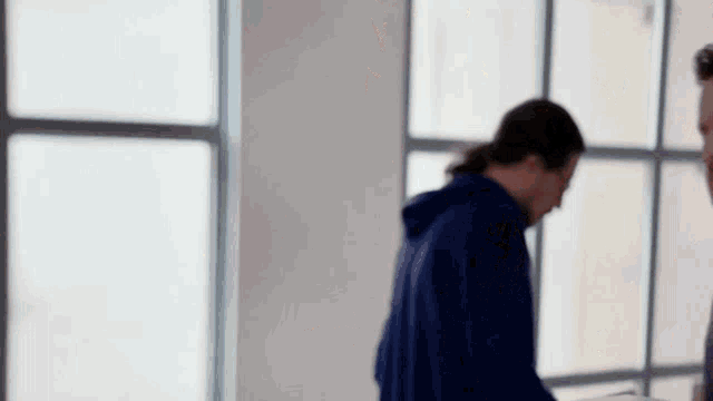 a man in a blue hoodie stands in front of a window talking to another man