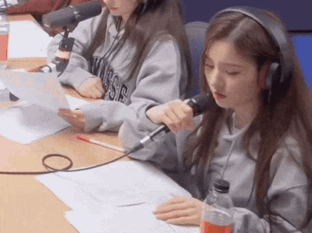 two girls wearing headphones are sitting at a table with microphones and a bottle of orange soda