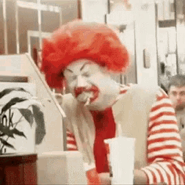 a clown in a red wig is sitting at a table in a restaurant eating a hamburger .
