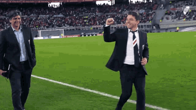 two men are walking on a soccer field and one of them is wearing a suit and tie