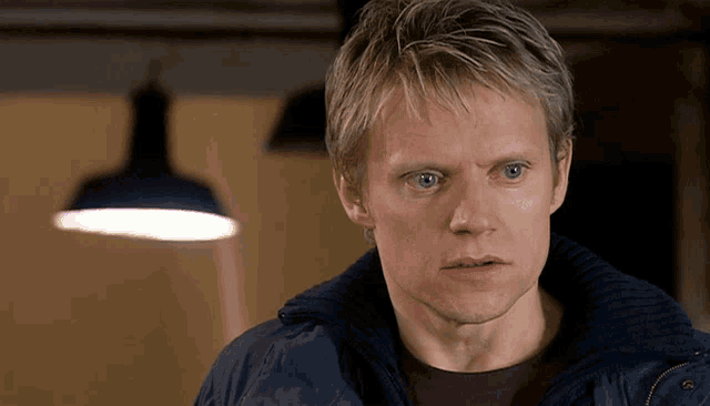 a man in a blue jacket looks at the camera with a lamp in the background