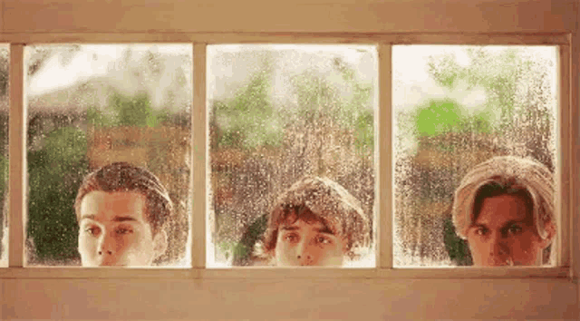 three young men are looking out of a window with rain drops on it .