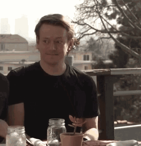 a man in a black shirt is sitting at a table with mason jars and a plant .