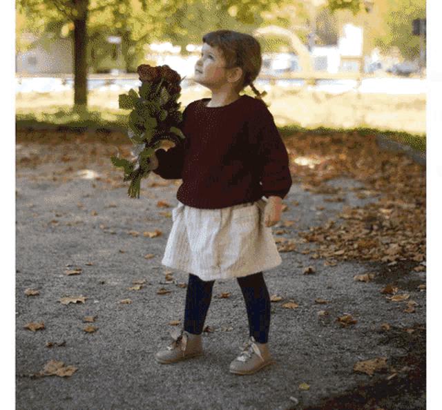 a little girl in a maroon sweater and white skirt is holding a bouquet of red roses