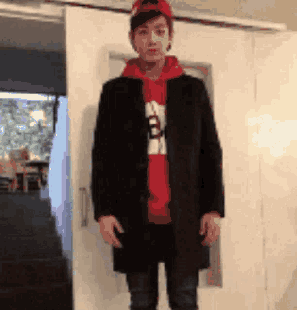 a man wearing a red hoodie and a black coat is standing in front of a white door