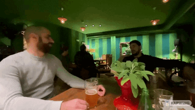 two men are sitting at a table with a plant and a glass of beer in front of them