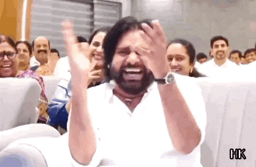 a man with a beard is sitting in front of a crowd and laughing with his hands in the air .