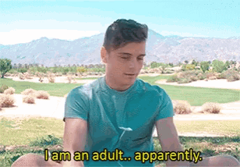 a man in a blue shirt is sitting on the grass and says i am an adult apparently